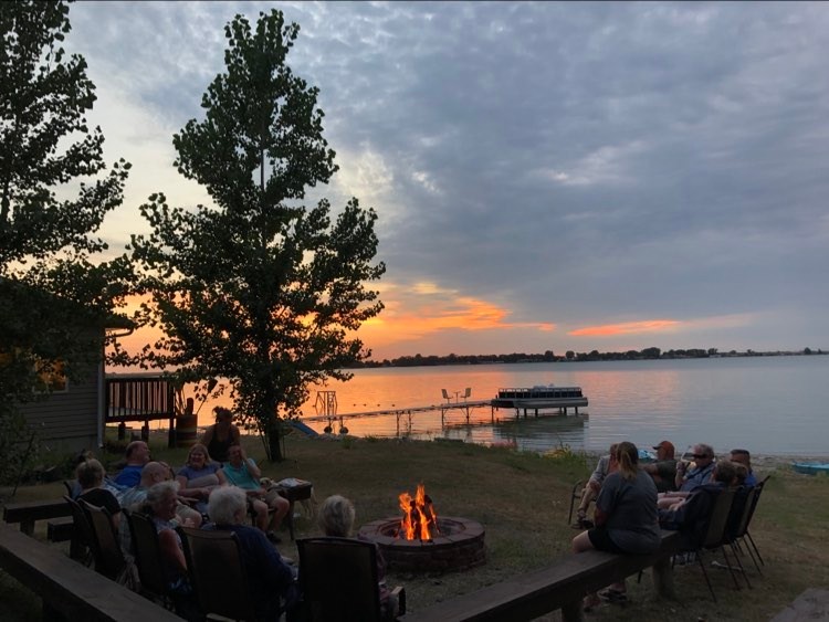 Photo is of Camp Grassick with campers around a roaring campfire with the sunset fading across the lake.