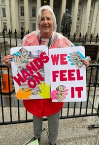 Zelda in front of the US Treasury following the March with her signs one which has “Hands On” and the other “We Feel It” in large red lettering and showing colorful hands holding clusters of bills. 