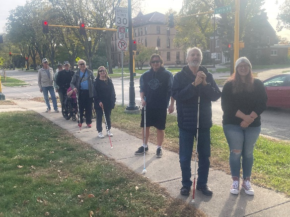 9 people walk and one person uses a wheelchair on a sidewalk along a street in autumn. Several people are using white canes to navigate.