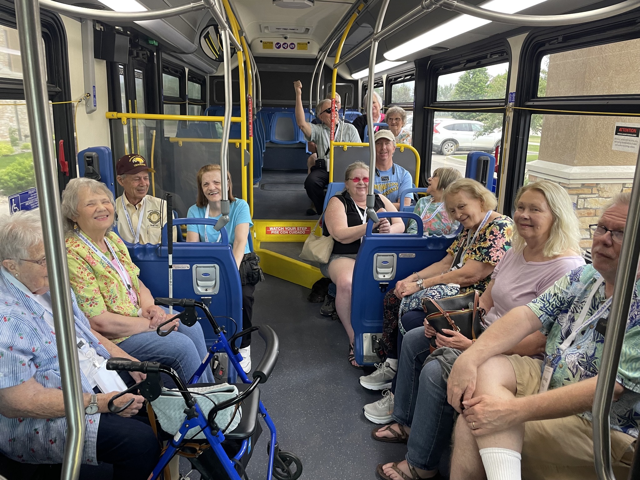 14 people sit on a city bus with yellow poles and blue details, many of them smiling for the photo.