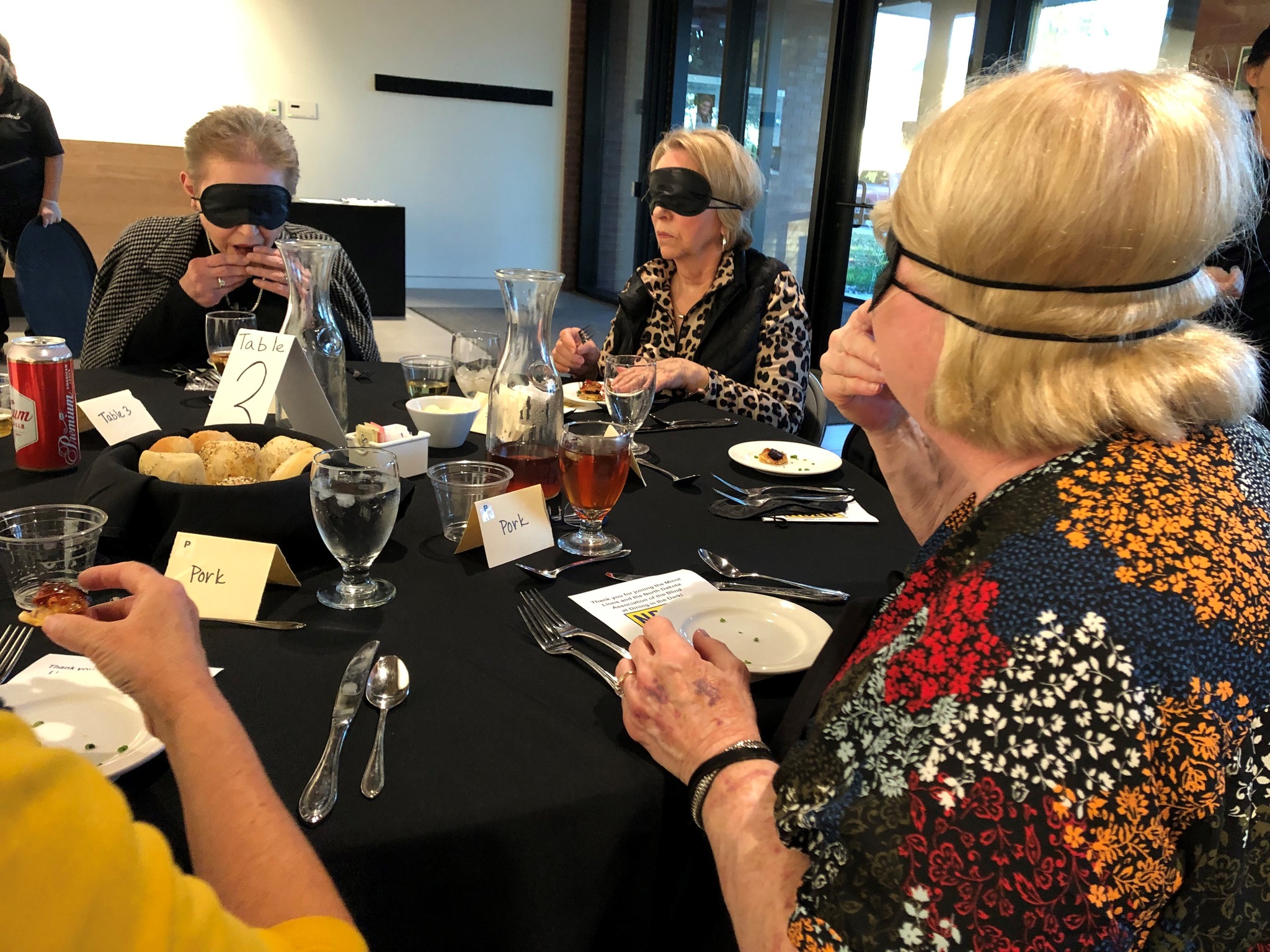 Three women wearing black blindfolds sit at a table covered with a black table cloth. They take bites of a small appetizer.