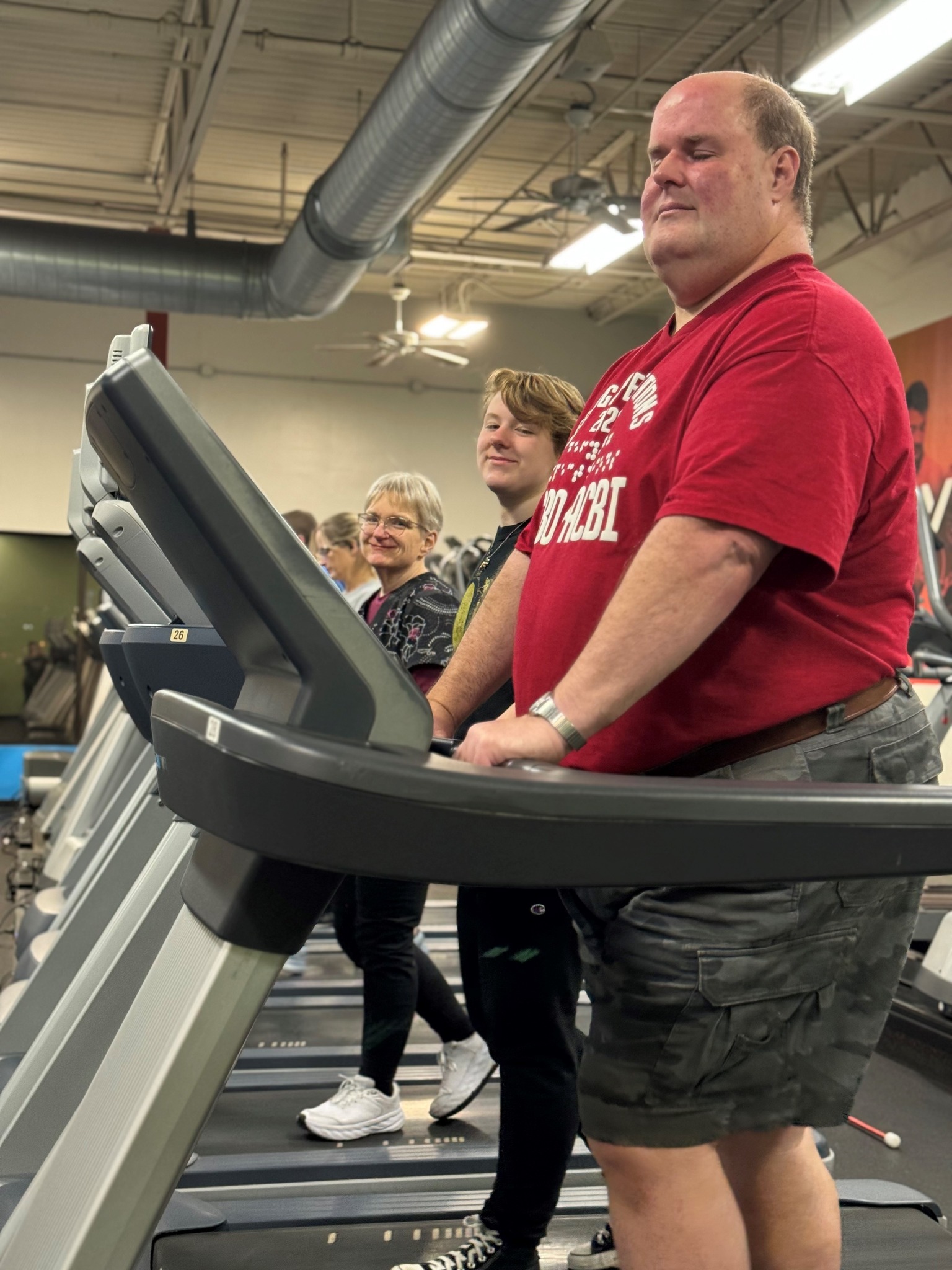 Brant wears a red T-shirt and khaki shorts. He smiles while on a treadmill with a teen and woman smiling behind him on their own treadmills.
