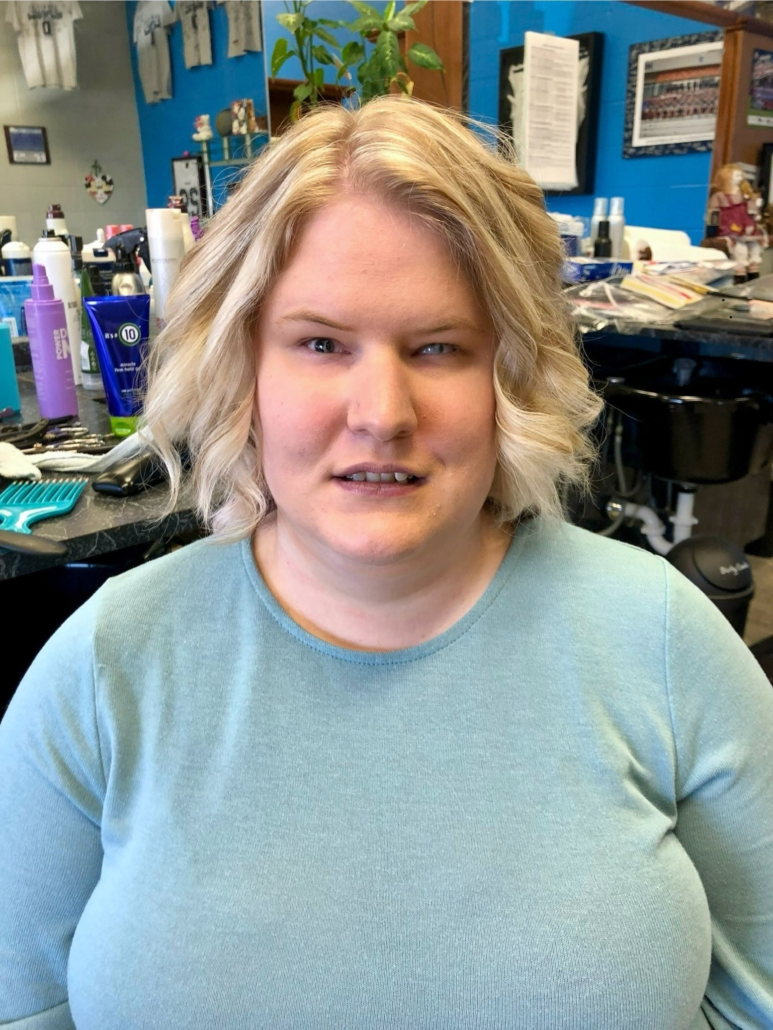 Lexee has curly, blonde hair and wears a light blue shirt in a salon.