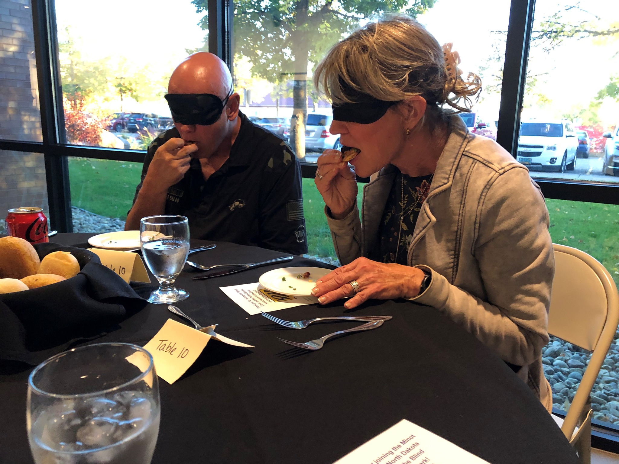 A bald man and woman wear blindfolds. They're both in the middle of taking a bite of food at a dining table. The sun is shining through the windows behind them.