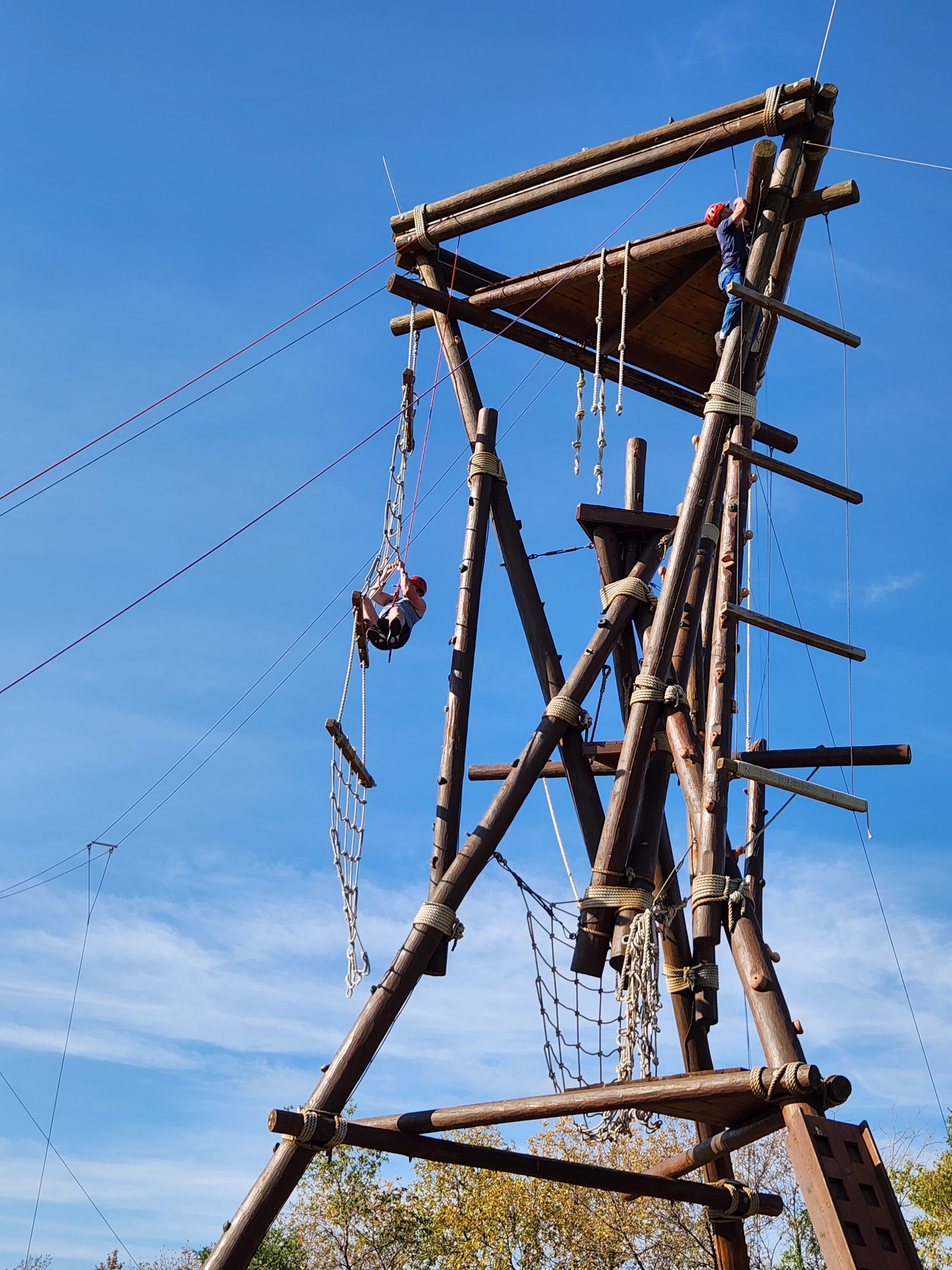 Two men climb a large, wood tower constructed of logs and rope. At left, one works his way up a rope ladder. At right, one nears the top of a log.