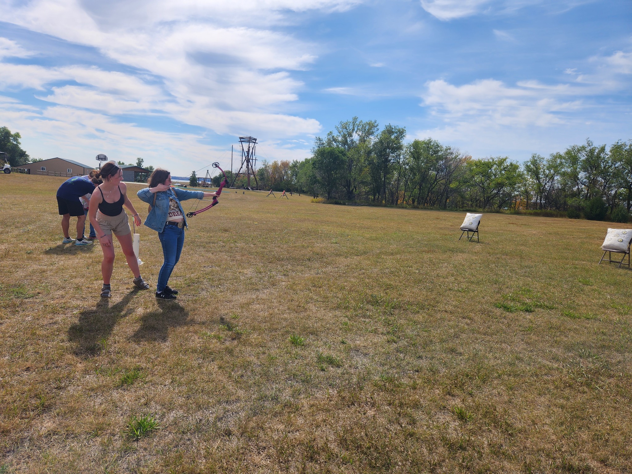 On a grassy field, a woman in a denim jacket and blue jeans pulls back on a bowstring. Another woman stands behind here, eyeing the targets on the far right.